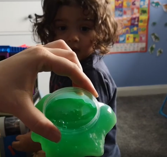 toddler in background while the opened slime container is being shown to the camera by mothers hand